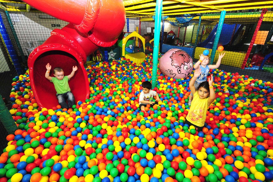 Three children standing in soft ball pit with another child just coming out of a red slide just about to land into the soft ball pit