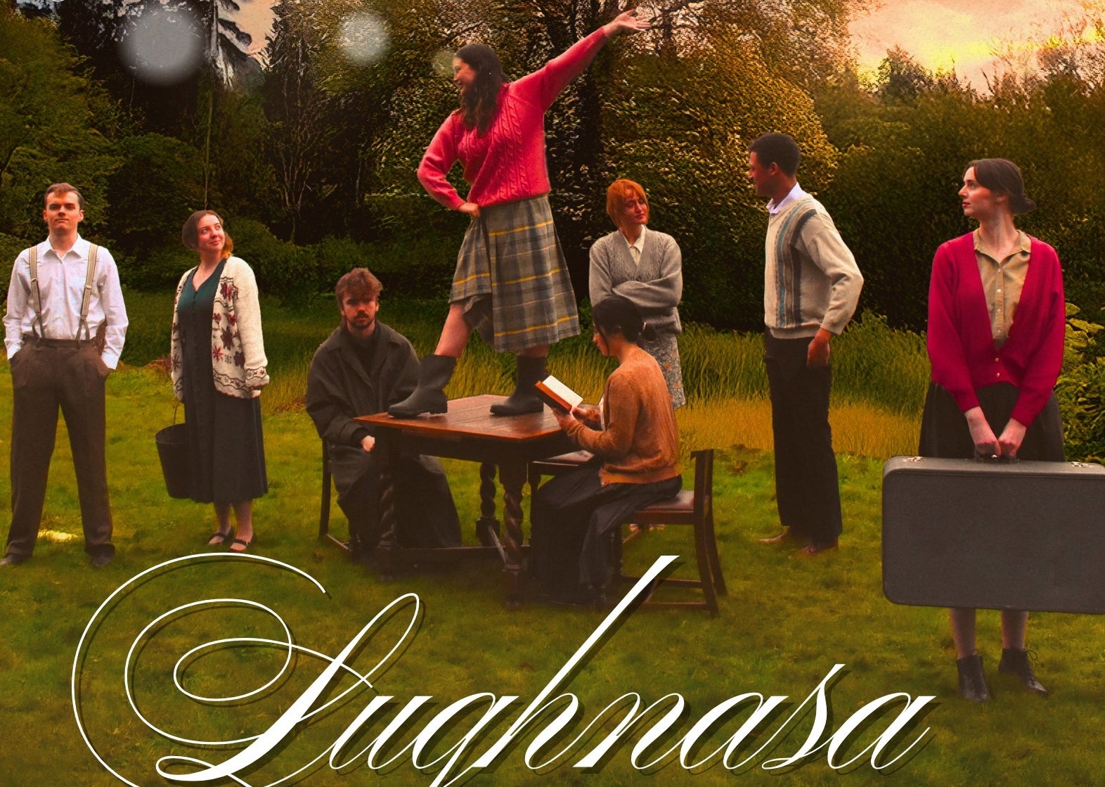 Dancing at Lughnasa - An Táin - A group of people standing outdoors on grass with 2 people seated at a table.