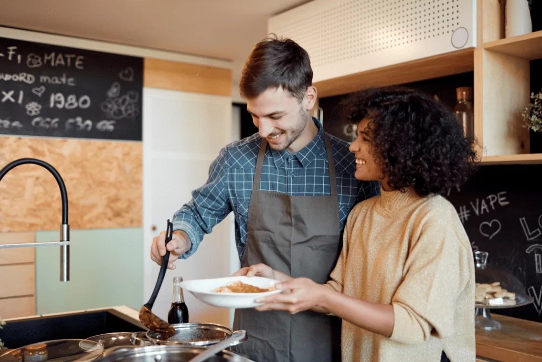 A couple are smiling, standing in a kitchen cooking.