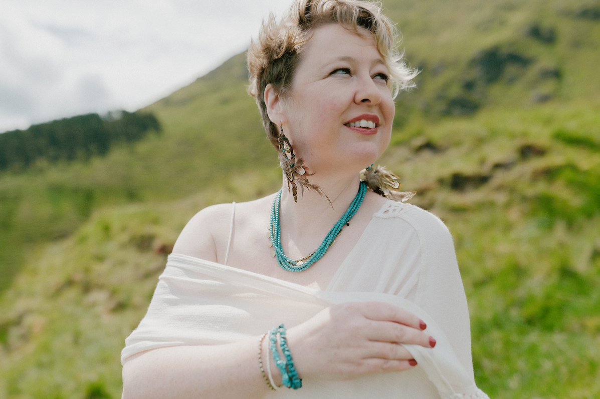Photo: Berit Alits. A woman is wrapping a white shawl around her shoulders looking away to her left, outdoors with grassy hill in the background.