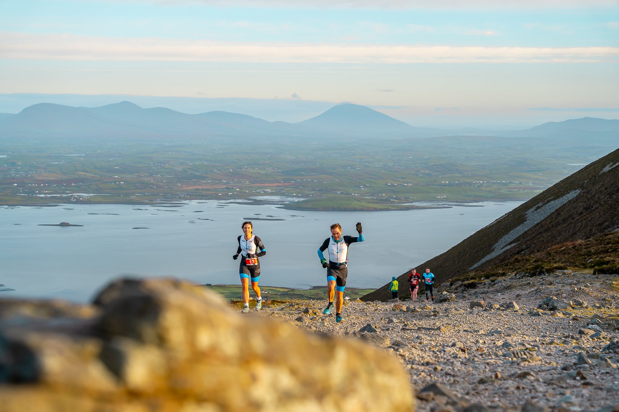 2 runners seen on a small rocky area with views of hills in the background.
