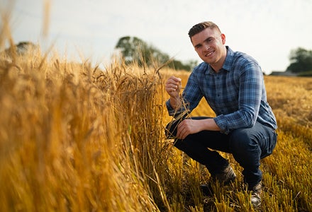 A smiling man crouched down beside golden wheat growing in a field.