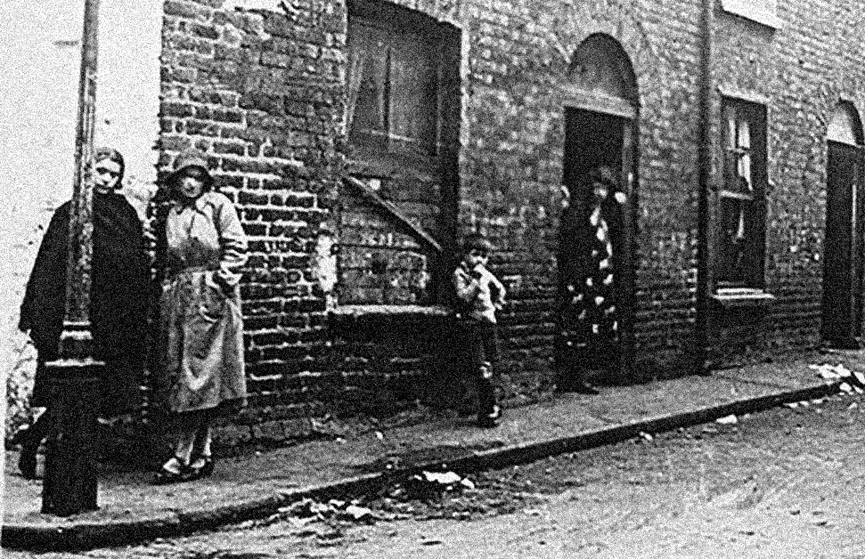 Very old black and white photo of 2 women standing at corner of row of old, brick terraced houses.
