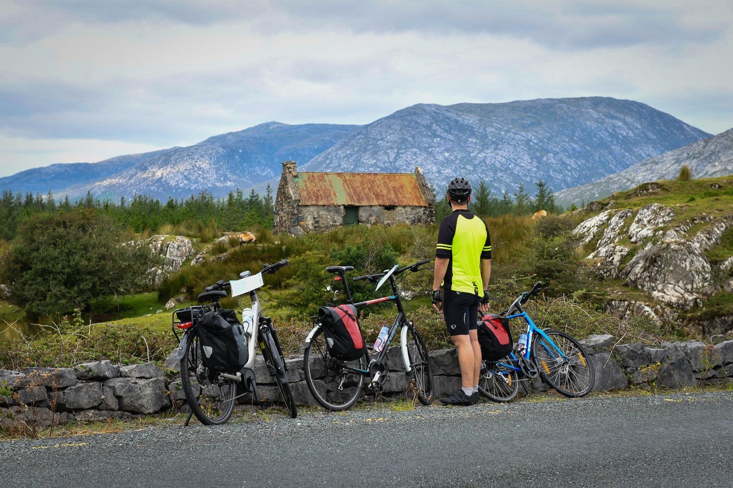 A man in cycling gear standing with bicycles at a stone wall looking at an old cottage
