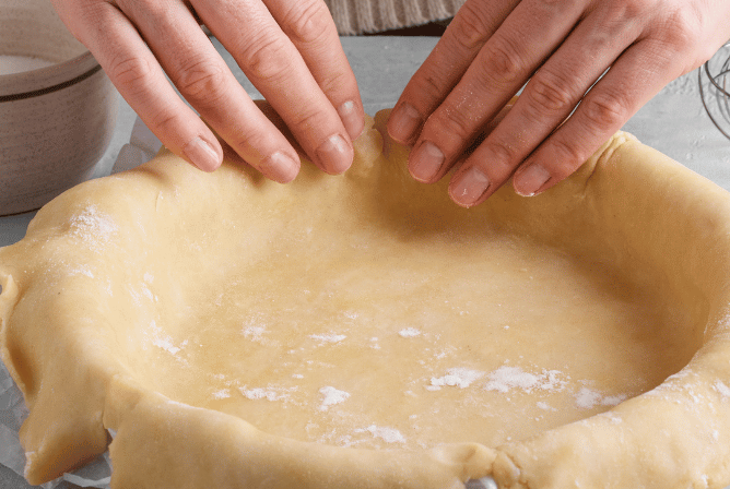 Fingers from 2 hands are touching the edge of a round dish covered with raw pastry.