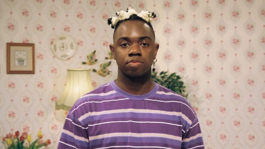 Cistin (the Irish word for kitchen) A man in a purple stripy T-shirt is looking directly ahead, against wall with old fashioned style wallpaper and ornaments.