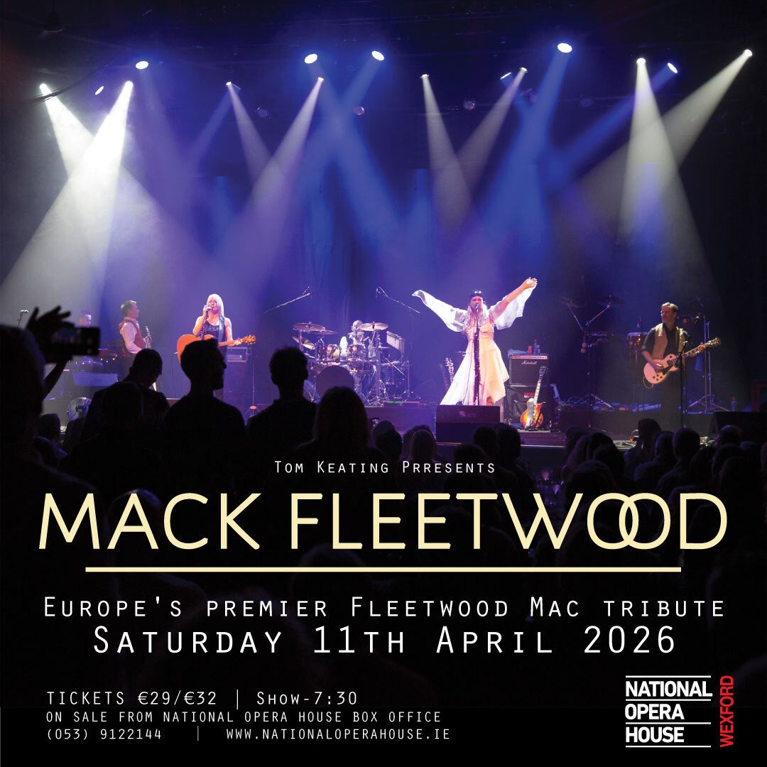 Distant view of a stage, with person dressed in white with arms outstretched and other band members, lit from above by many downward spotlights