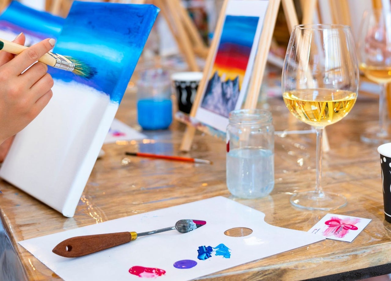 A person painting a canvas at a table with brushes and a glass of wine