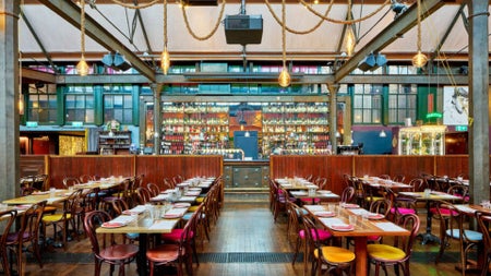 Restaurant interior with high industrial ceilings hanging lights wooden tables and a central bar area lined with bottles
