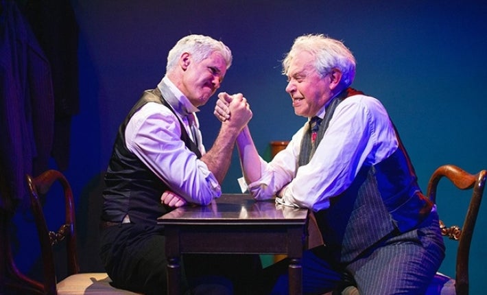 2 men in shirts and waistcoats arm wrestling on a small table.