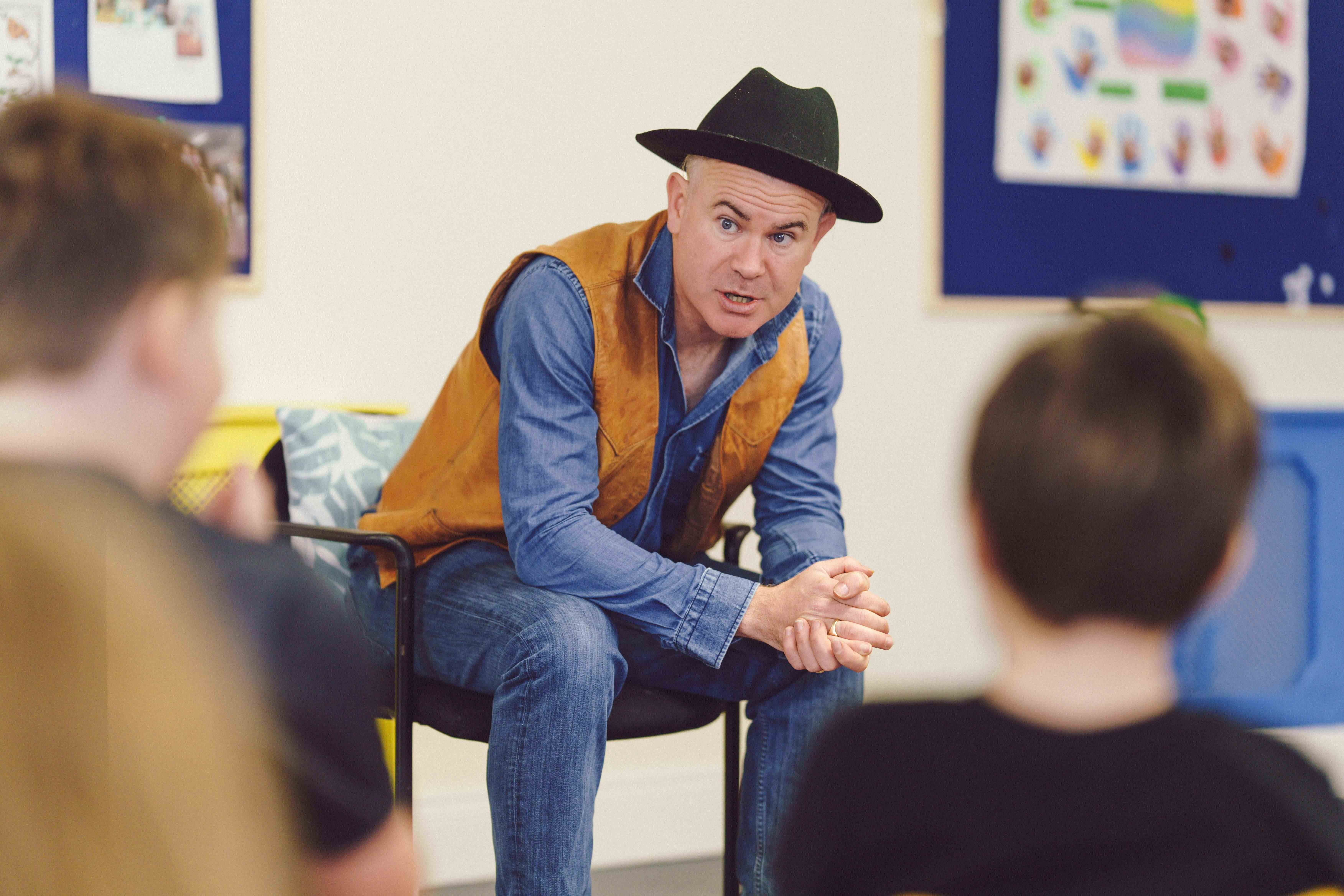 Killian Storytelling for Children - a man in a black hat, seated leaning forward talking, blurred rear view of children in foreground.