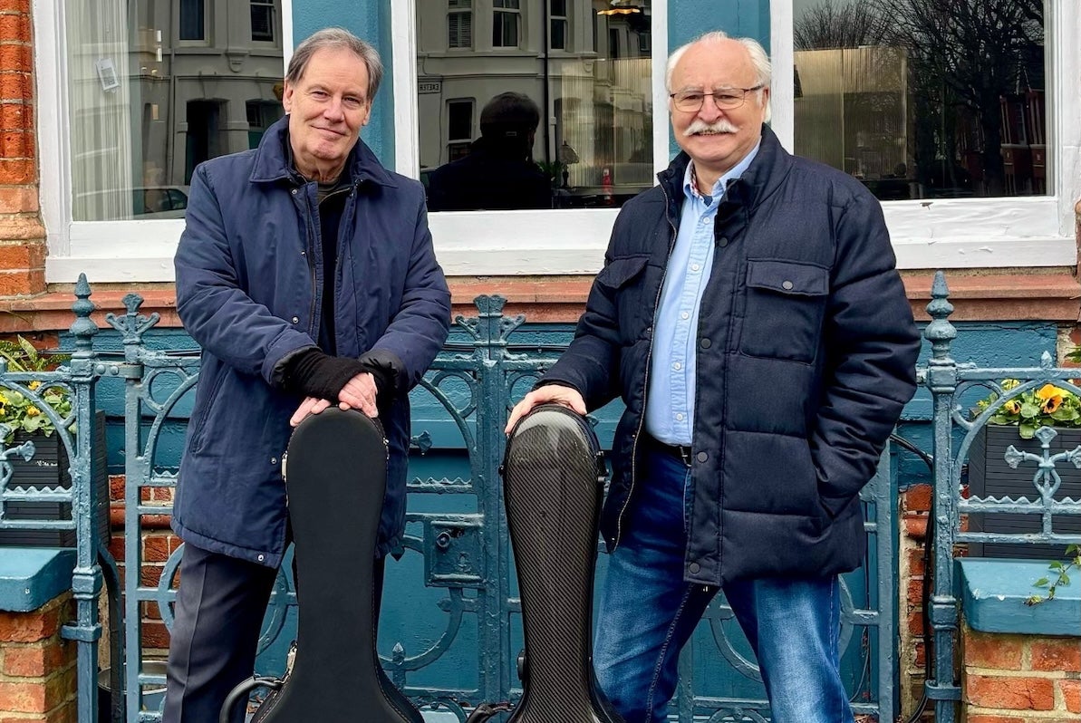 2 older smiling men with guitar cases standing in front of railings.