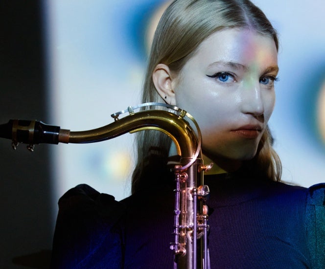 A serious looking woman in dark top with part of saxophone visible she is holding in front of her.
