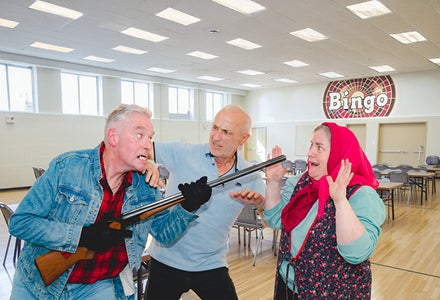 A man is holding a rifle at a horrified woman in a headscarf with another man holding him back in a hall with table and chairs.