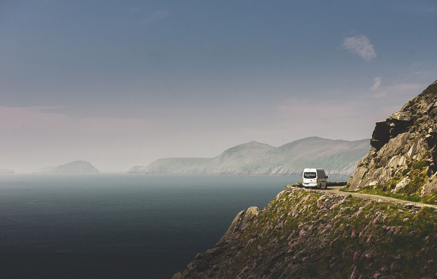 A bus with a logo parked along a cliff road with a dramatic coastal view