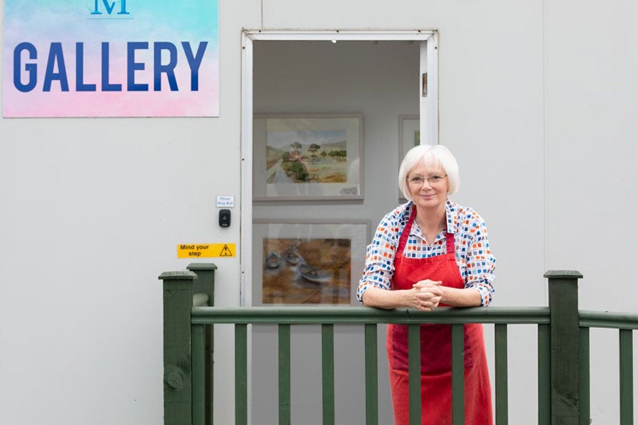 A lady leaning on a wooden fence with an open door behind her where there are pictures on a wall