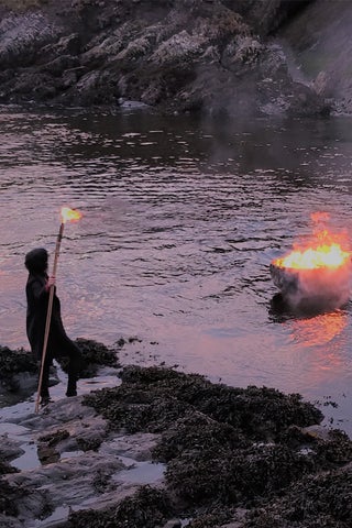 A person in a cloak holds a lit torch, watching a pire blaze in the middle of a misty river.