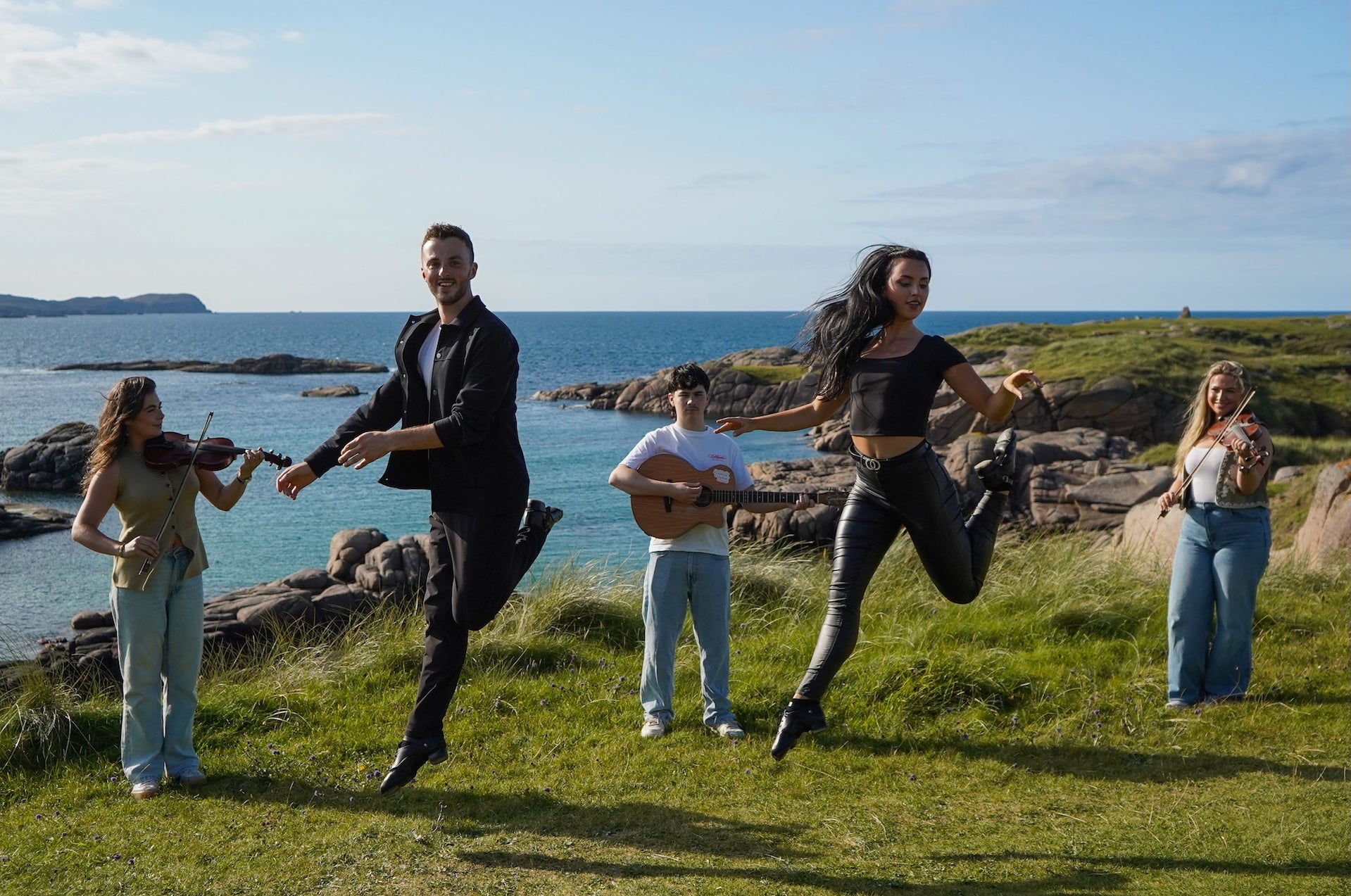 5 people standing with instruments or leaping in the air, outdoors on a rocky edge overlooking the sea.