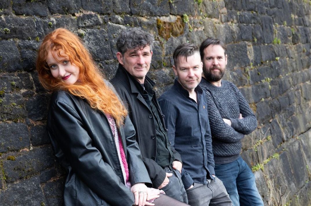 Anglo/Irish folk band The Haar. 4 smiling people leaning against a dark grey stone wall.