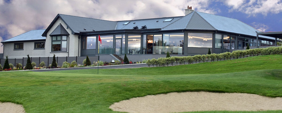 External view of the Clubhouse showing a low rise glass fronted building and overlooking bunkers on the green with a flagstick also visible on the green