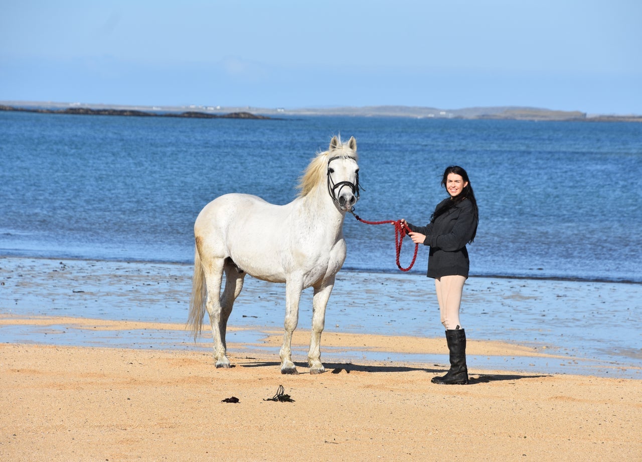 A woman in riding gear standing on a beach with a horse