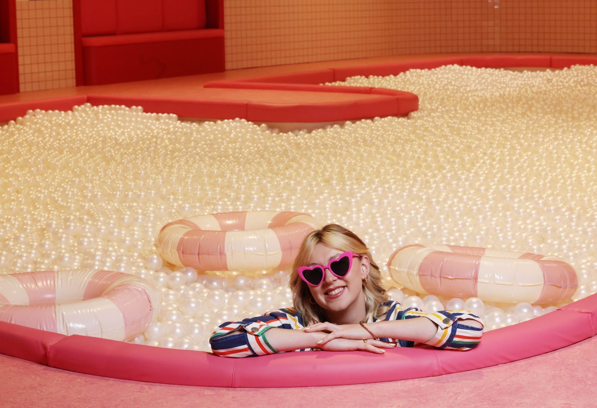 Person wearing pink heart shaped sunglasses in a full ball pit with three inflatable lifebuoys