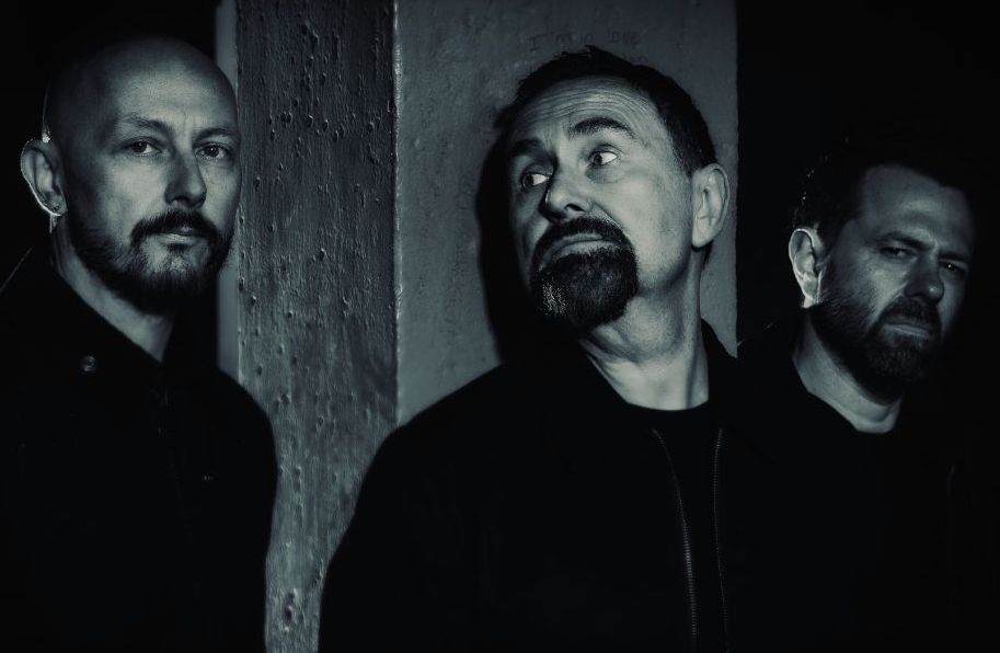 Darkly lit black and white photo of 3 men with short beards looking serious against dark, rough backdrop.