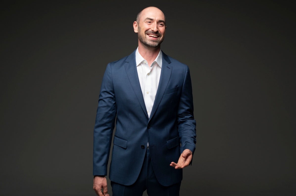 A laughing man in dark blue suit and open white shirt against plain dark background