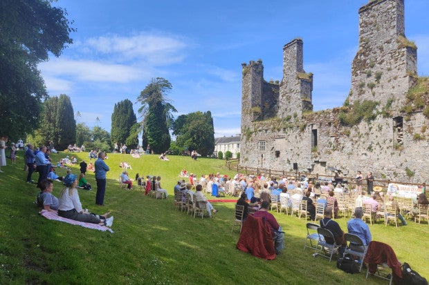 Free Open Air Recital Castlemartyr, people sat in chairs spread across a sloping green area looking at group performing against background of ruined castle.