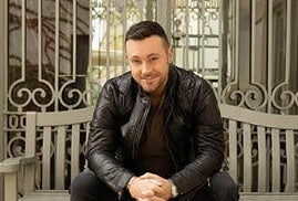 A smiling man dressed in black seated on a bench in front of metal railings