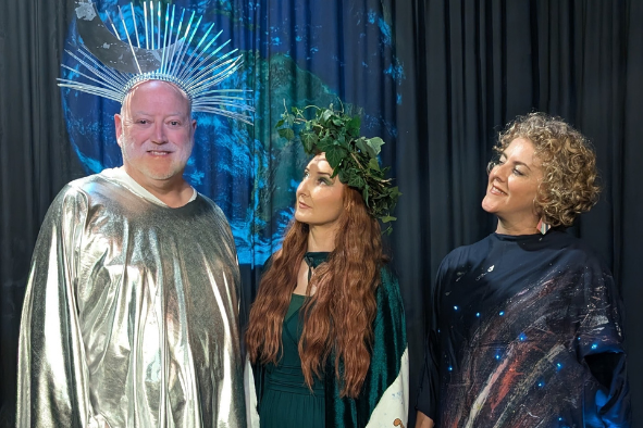 2 women are looking at a smiling man in a gold smock with a silver headpiece