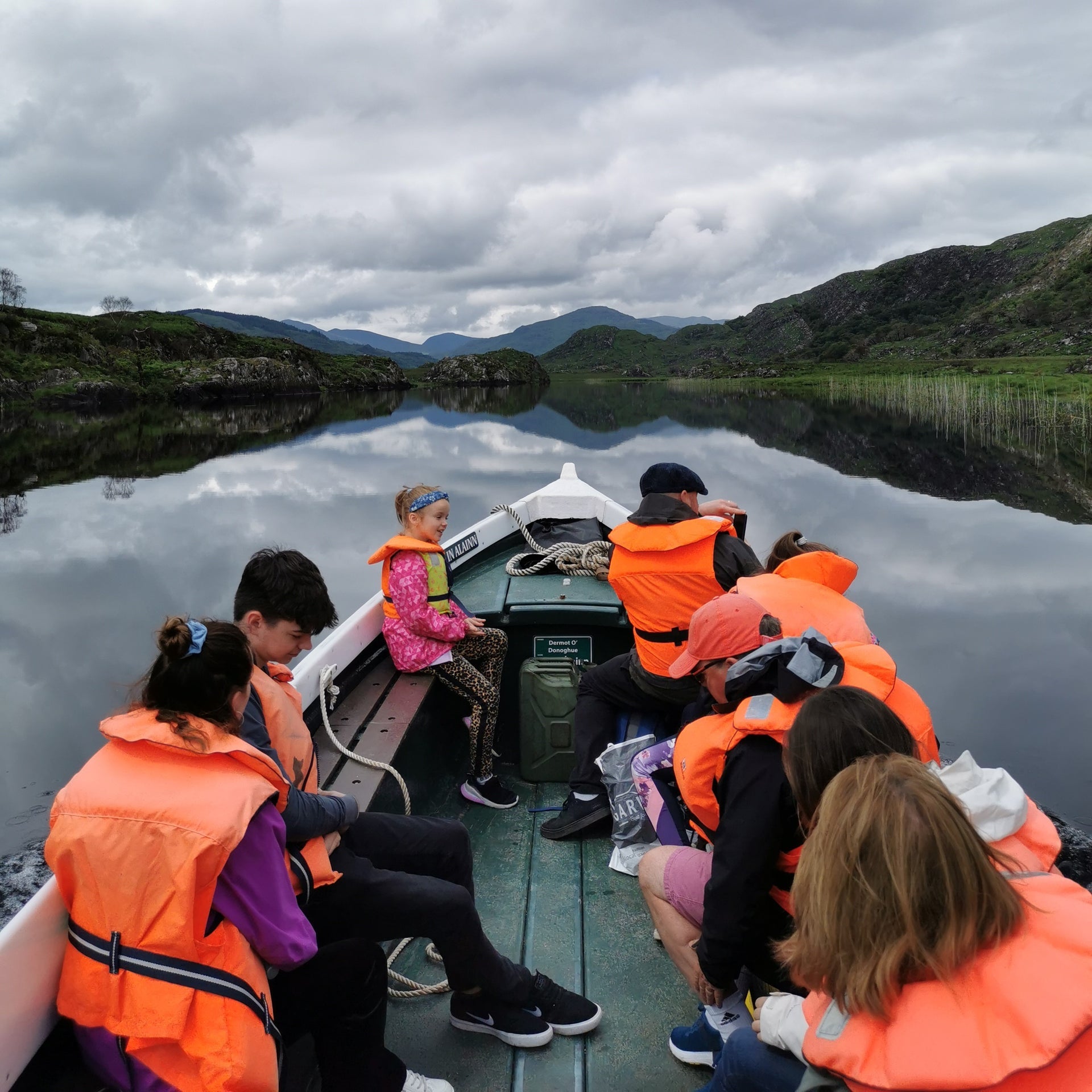 Group of people wearing orange life jackets sitting on a boat sailing on a lake with mountains in the background