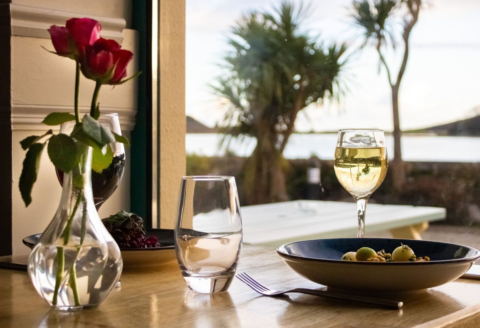 Table with food a rose in a vase and two glasses on it and views of some trees and the sea