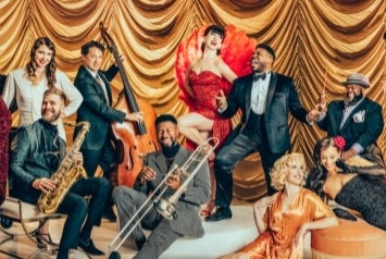 A group of musicians with instruments in vintage suits and dresses, some seated or standing or laying on a light coloured carpet.