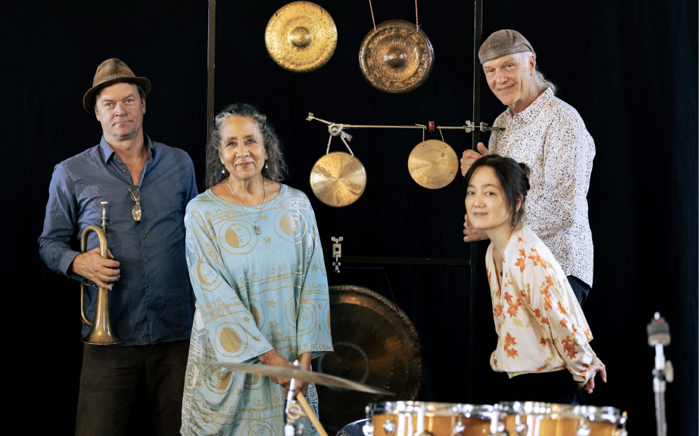Photo by Patrician Pereira of 4 smiling people with percussion instruments including large drum and various round cymbals hanging on a display against black background.