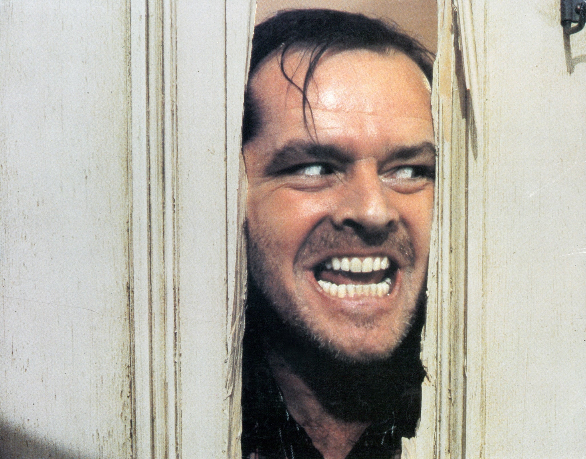 A man with crazed expression is looking through a hole in a door.