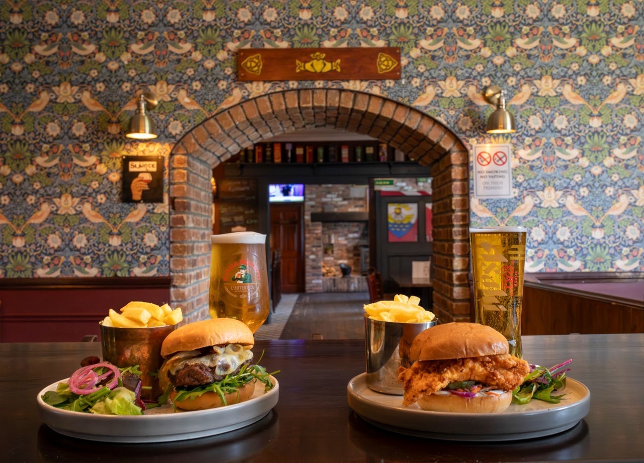 Two plates of burgers and chips with drinks on a bar counter framed by a patterned wallpaper and brick archway