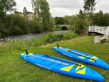 Two blue stand up paddleboards lie flat on a river bank near an old bridge