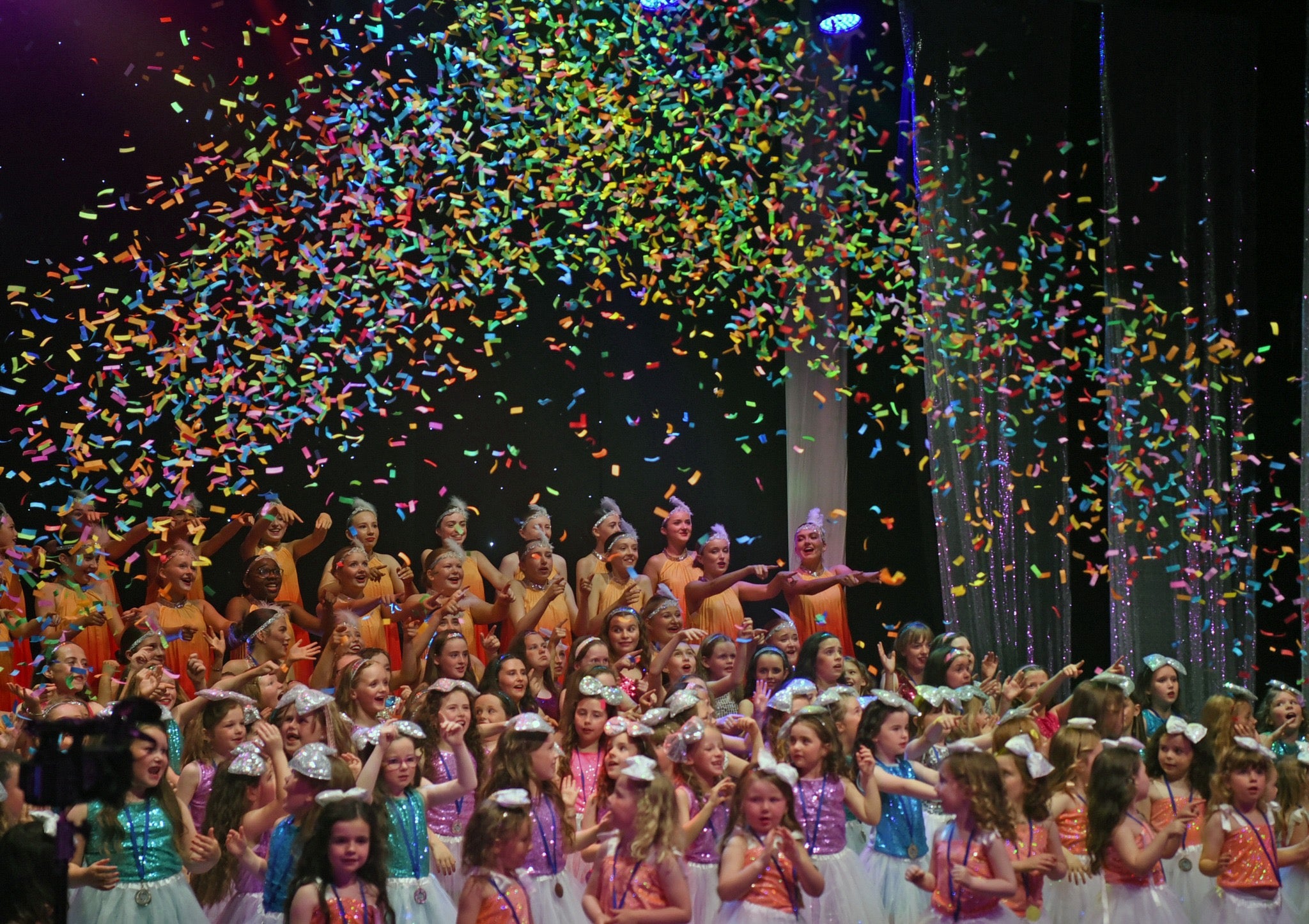 A large group of children gathered on a stage with confetti falling from above