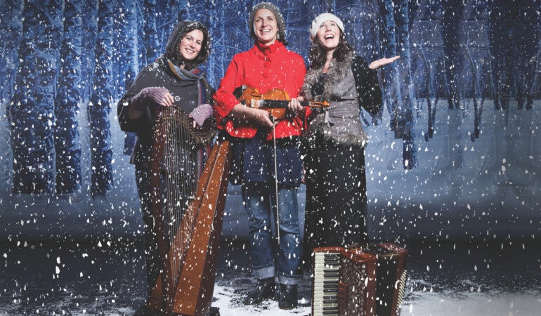 3 smiling women standing with musical instruments in snowy scene
