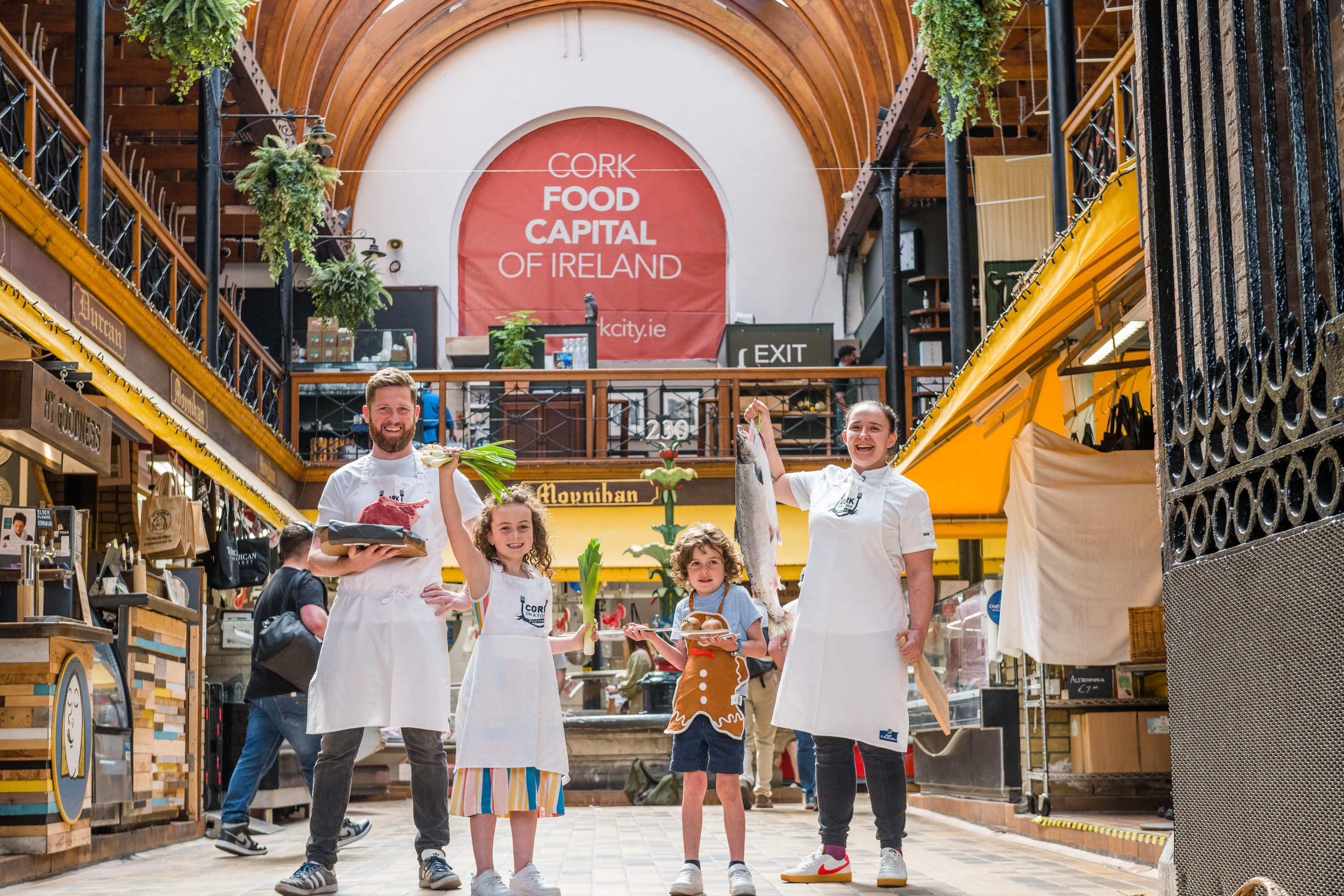 Group standing inside a market hall. One adult holds a tray of raw meat, a child holds green onions, another child holds baked goods, and one adult holds a large fish. A banner reading “CORK FOOD CAPITAL OF IRELAND” hangs above.