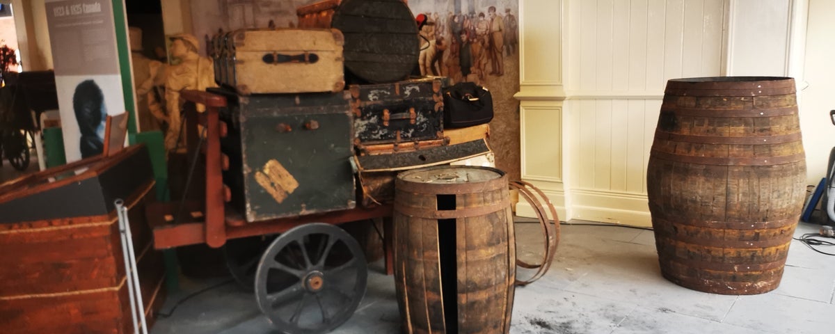 A heritage display of old luggage on a cart ready for loading on a ship with two barrels nearby