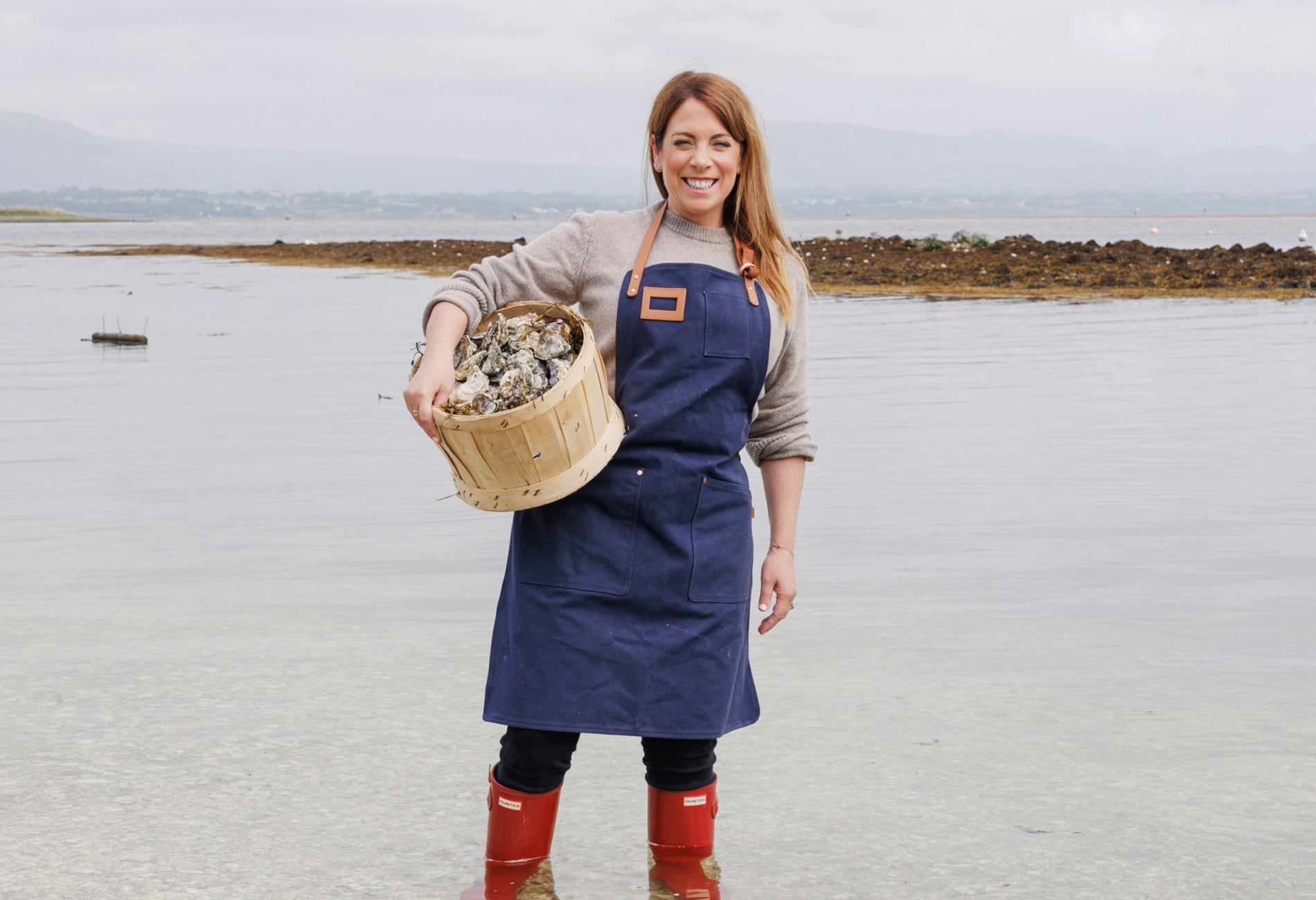 Person standing in a grey sea holding a wooden bucket of oysters