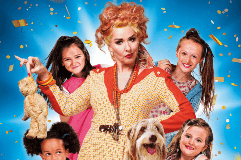Claire Sweeney as Miss Hannigan in Annie, a woman with rye expression surrounded by 4 young girls and a dog.