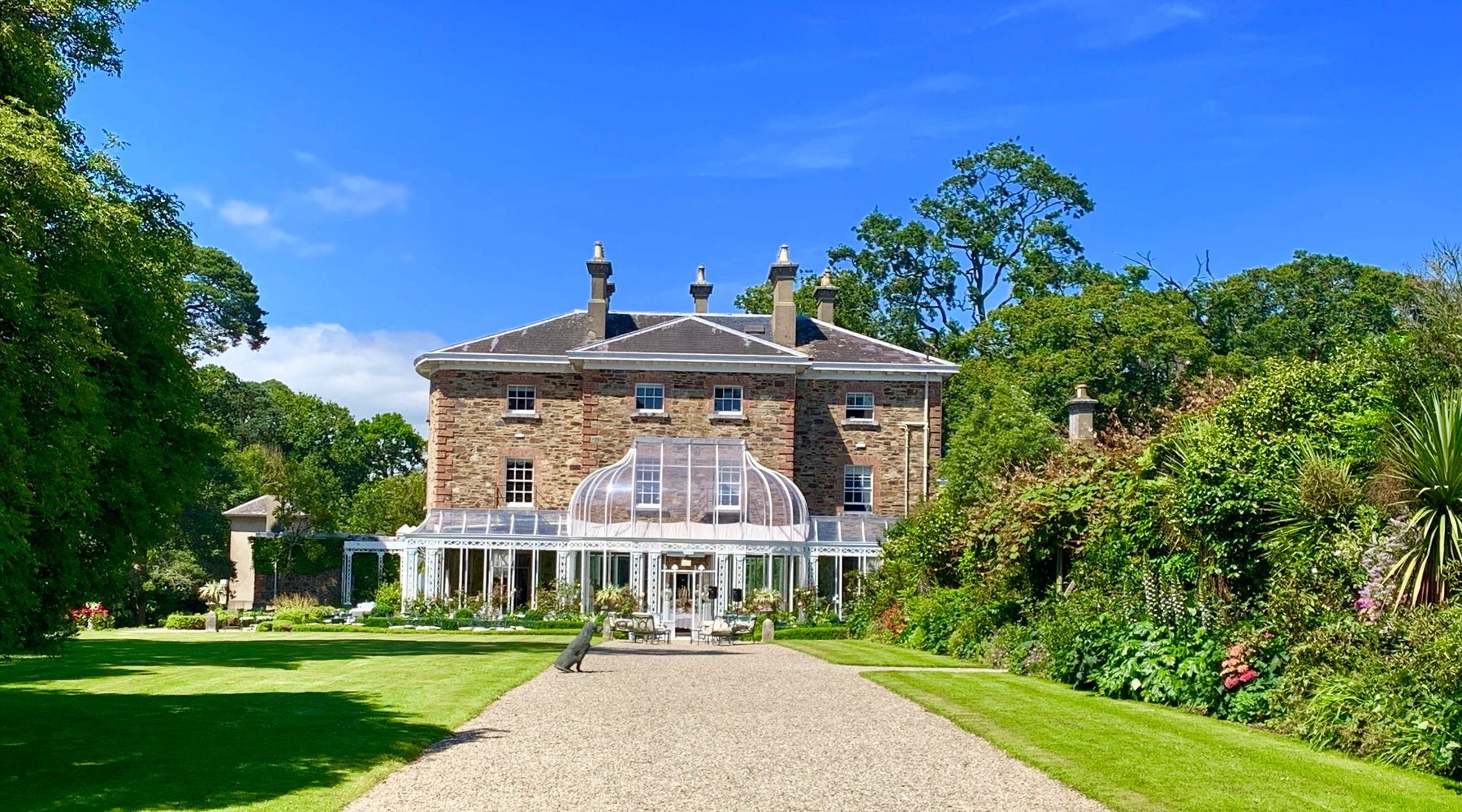 A large period house with a glass house at the front surrounded by trees and a lawn