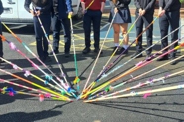 View of lower half of teenagers all holding long, thin, decorated poles into a central point on a playground.
