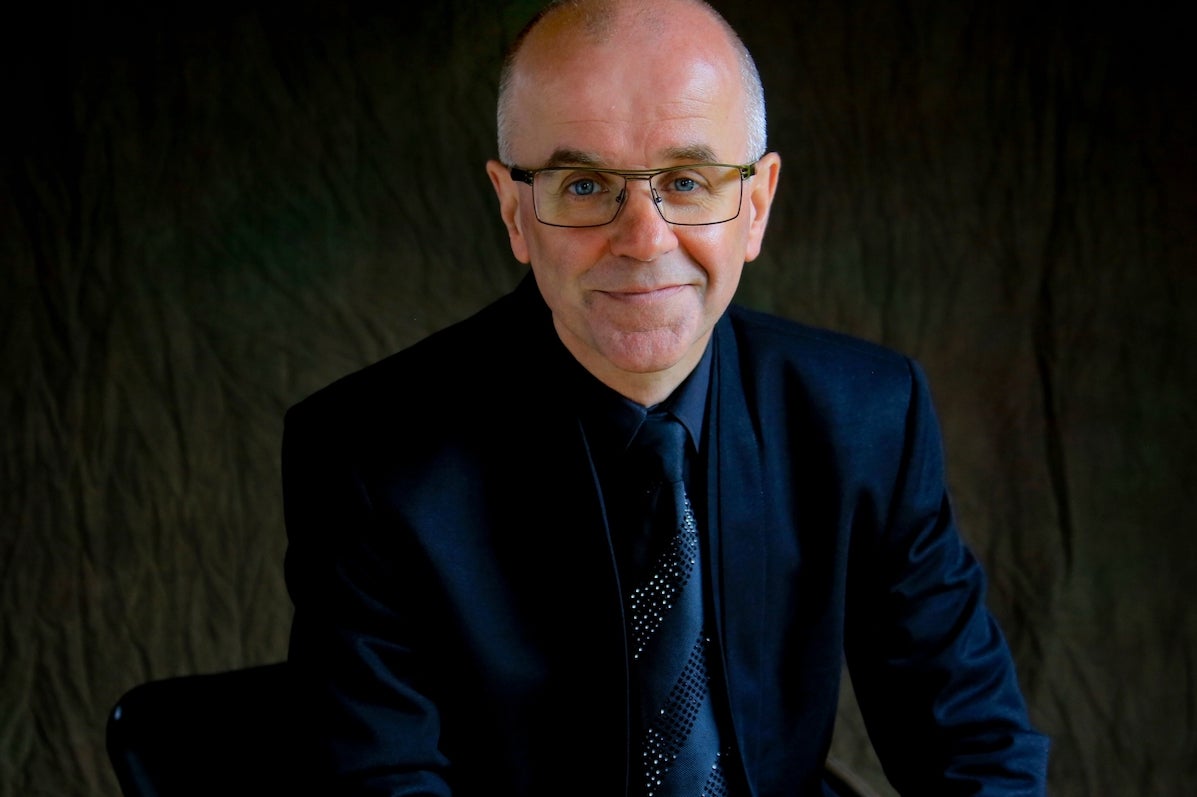A smiling man in dark suit, tie and jacket against a dark, plain background.