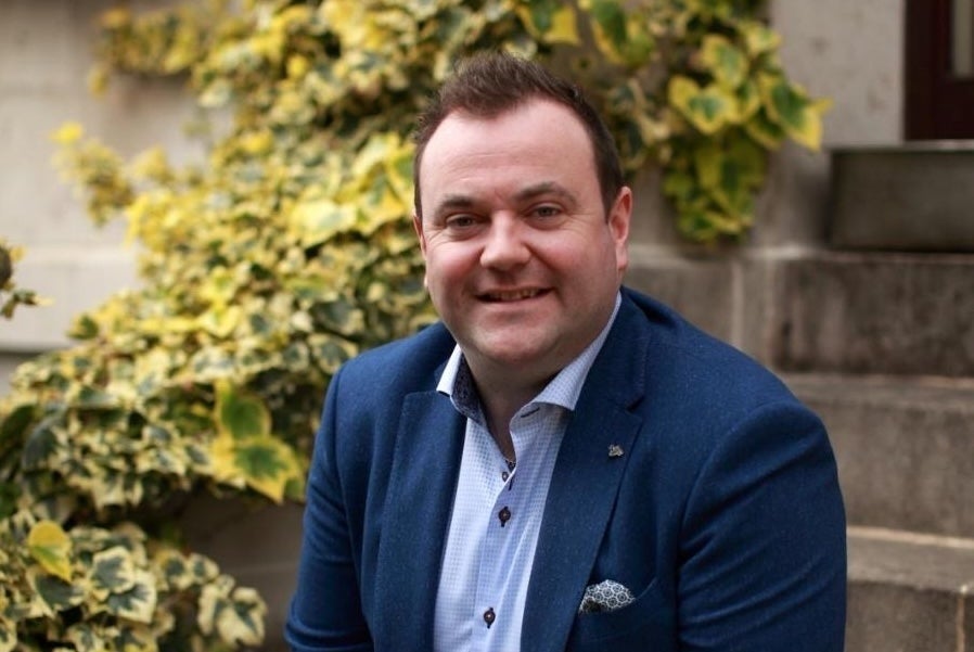 A smiling man in navy jacket seated on stone steps outside with bushes beside