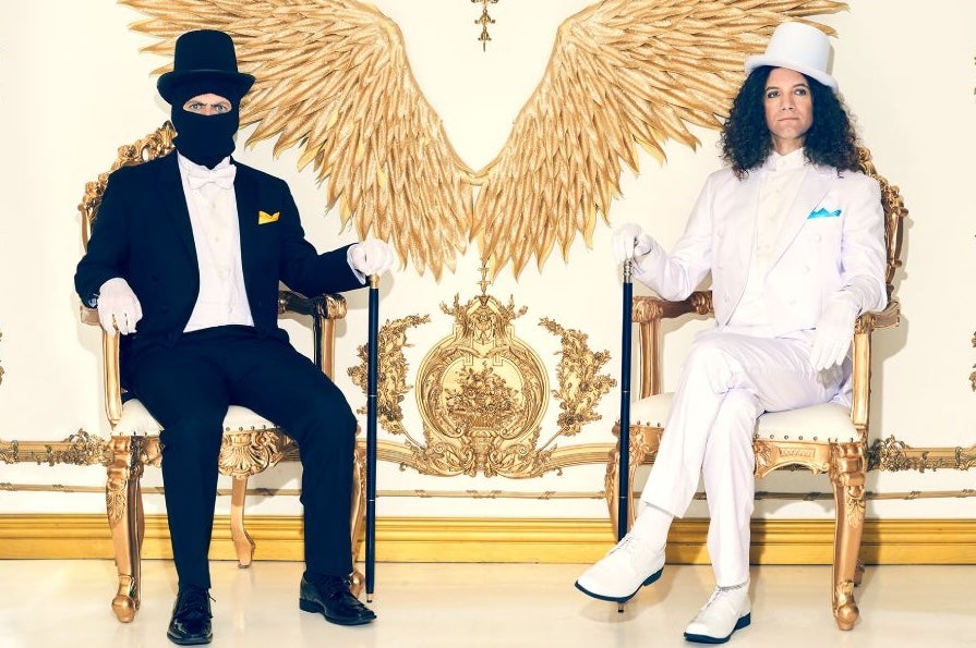 2 men seated in ornate gold chairs, one in black suit with face covered by a mask, the other in a white suit, against ornate white and old decorated wall.