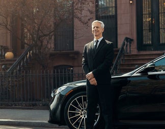 Person in a dark suit standing by a car on a street with steps in the background